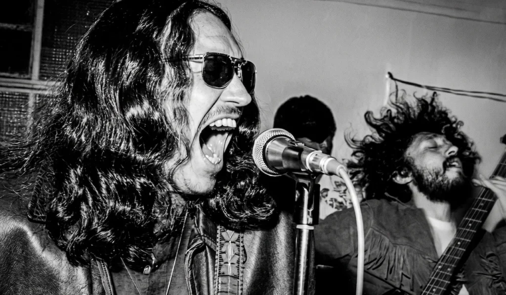 Vocalista de la banda mexicana Enigma! cantando en vivo con gafas de sol y chaqueta de cuero, escena del rock nacional de los años 70 en blanco y negro.