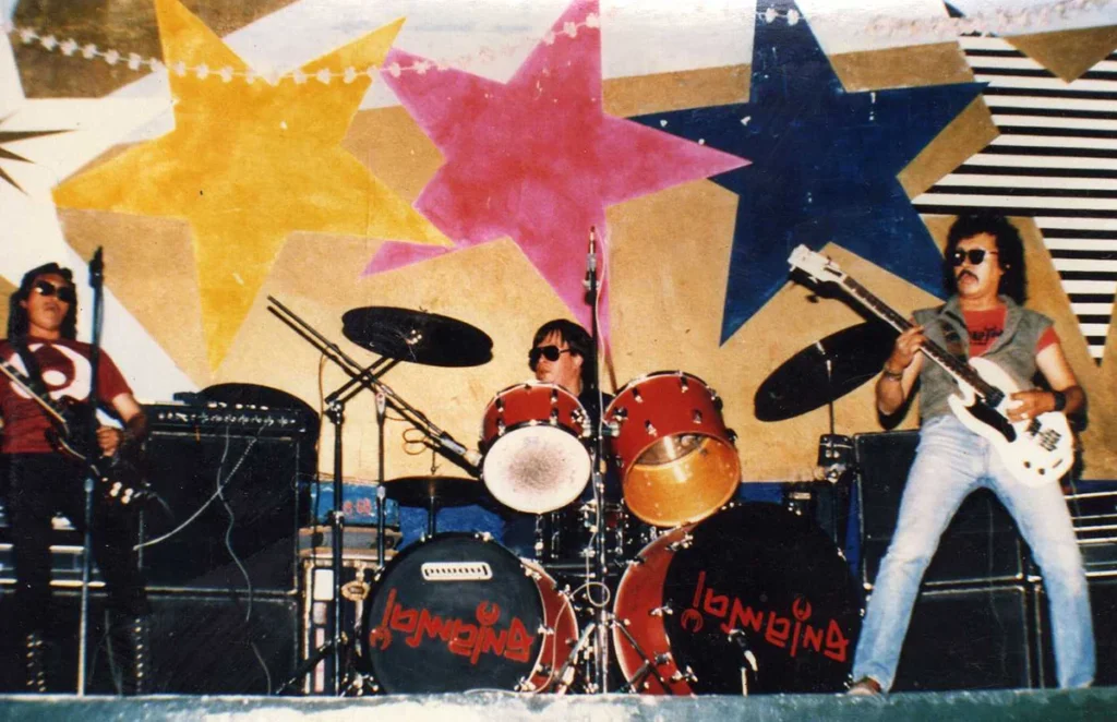 La banda de rock pesado Enigma en el escenario, destacando la batería roja con el logotipo clásico del grupo y los integrantes usando gafas de sol frente a un fondo de estrellas coloridas.
