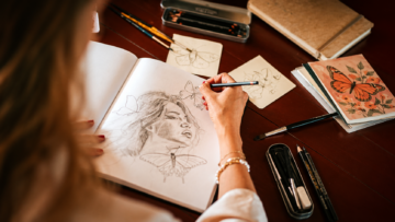Vista desde el hombro de la artista Nicolette van den Hadelkamp dibujando a lápiz un retrato femenino rodeado de mariposas en su cuaderno de bocetos.