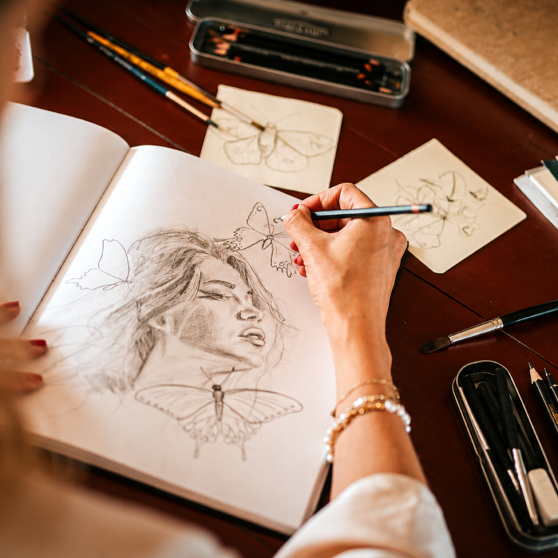 Vista desde el hombro de la artista Nicolette van den Hadelkamp dibujando a lápiz un retrato femenino rodeado de mariposas en su cuaderno de bocetos.