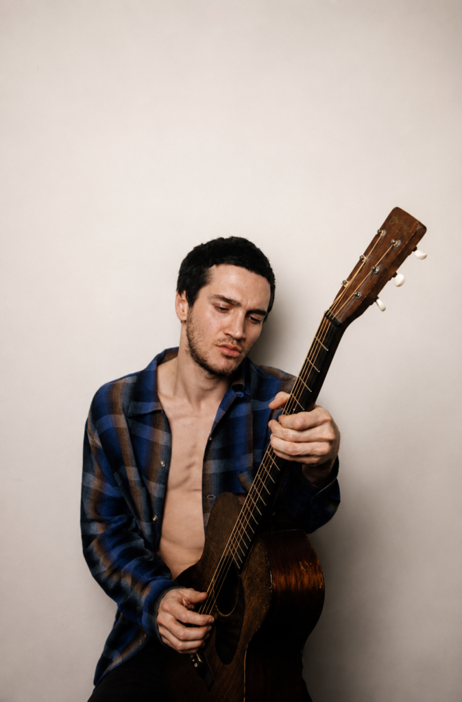 El músico John Frusciante sin camiseta y usando una camisa de cuadros abierta, tocando una guitarra acústica vintage en una sesión de fotos del año 2000.