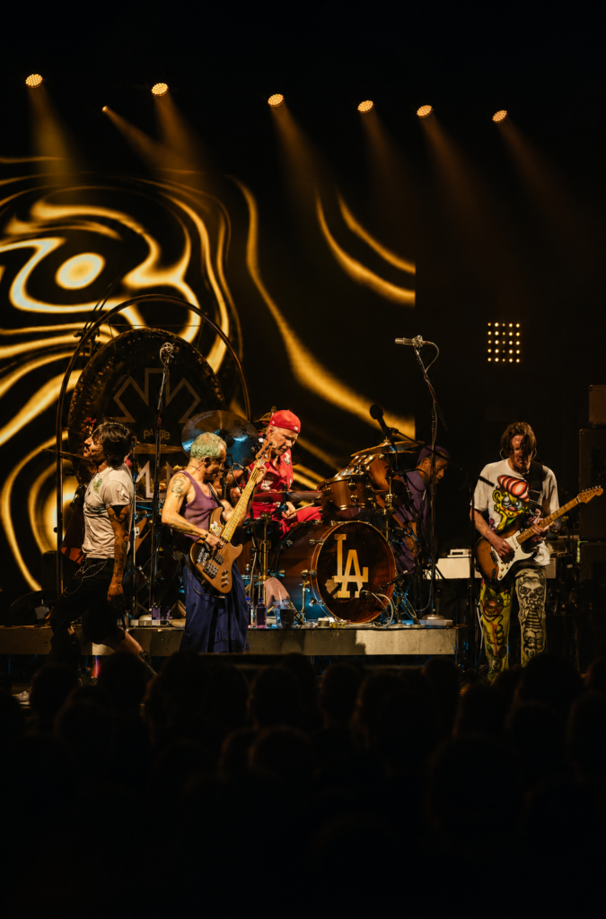 La banda Red Hot Chili Peppers tocando en vivo en un escenario iluminado en 2024, con el guitarrista John Frusciante tocando a la derecha y Flea al centro.