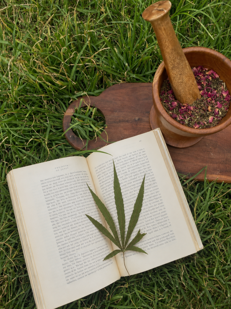 Una hoja fresca de cannabis sativa extendida sobre las páginas abiertas del libro "The Book of Japan", junto a un mortero de madera lleno de flores secas de lavanda y pétalos de rosa en el césped.