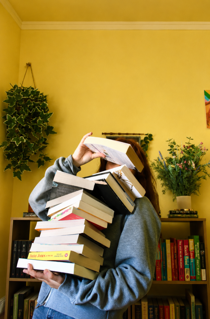 Joven con sudadera gris sosteniendo una gran pila inestable de libros frente a su cara, con una estantería llena y una pared de fondo de color amarillo brillante.