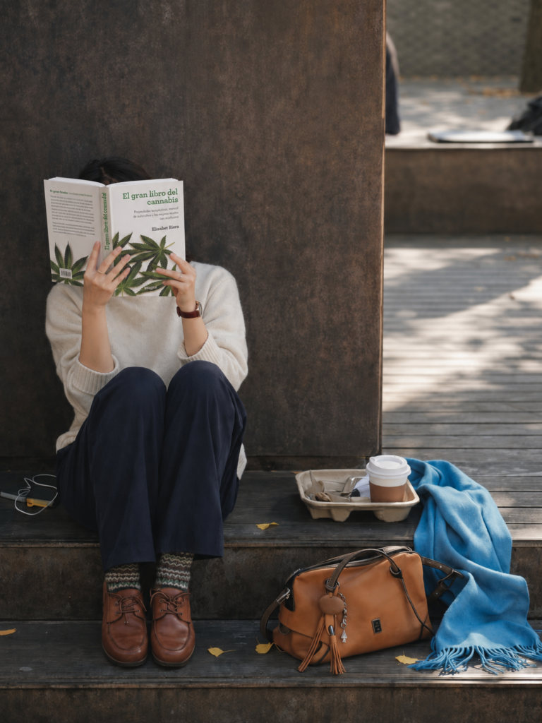 joven sentada en unos escalones de madera cubriendo su rostro con el libro "El gran libro del cannabis" de Elisabet Riera, junto a un café para llevar, un bolso de cuero marrón y una bufanda azul.