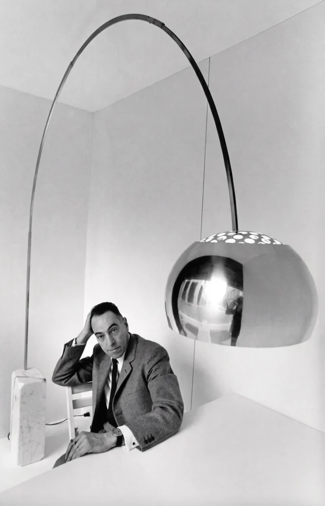 El diseñador Achille Castiglioni posando con la lámpara Arco de 1962, destacando la base de mármol de Carrara y el icónico brazo de acero inoxidable.