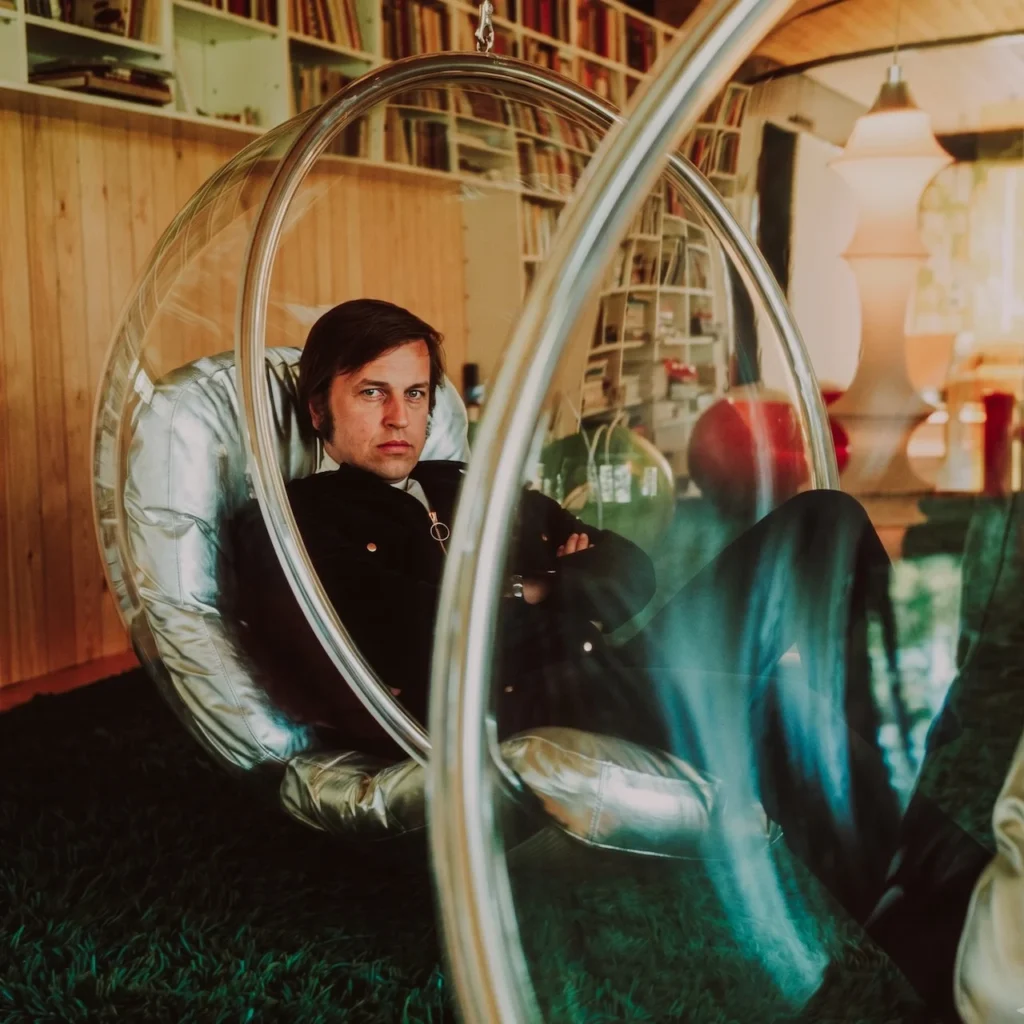 El diseñador finlandés Eero Aarnio sentado en su icónica Bubble Chair (Silla Burbuja) de 1968, destacando su estructura acrílica transparente y diseño suspendido de la era espacial.