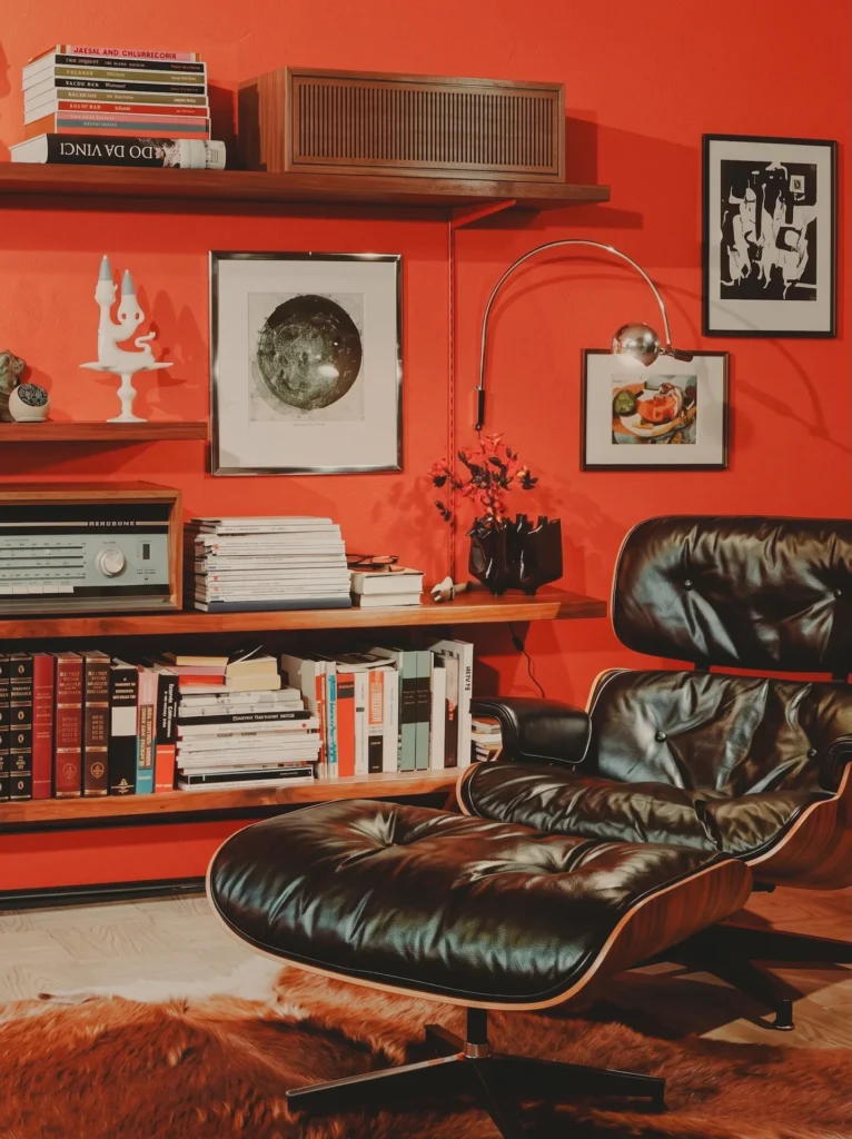 Silla Eames Lounge Chair con otomana en un estudio con paredes rojas, ejemplificando la apoteosis de la carcasa individual y la sofisticación del diseño industrial de 1950.