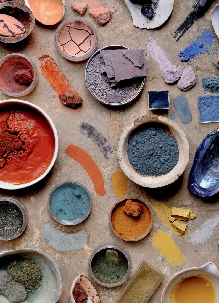 Vista cenital de una colección de pigmentos naturales, tierras y ocres minerales en recipientes de barro, conchas marinas y frascos, mostrando tonos rojos, azules, amarillos y verdes para la fabricación de pintura orgánica.