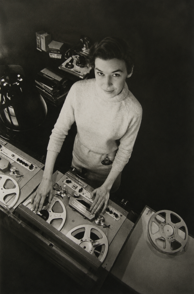 Fotografía en blanco y negro desde un ángulo cenital de la compositora Delia Derbyshire operando grabadoras de cinta de bobina abierta en el estudio del BBC Radiophonic Workshop.