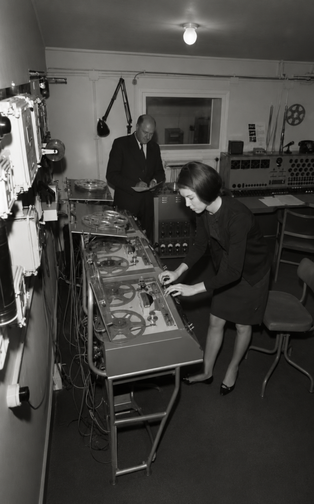 Fotografía en blanco y negro de la compositora Delia Derbyshire operando un banco de grabadoras de cinta magnética de bobina abierta frente al director Desmond Briscoe en el BBC Radiophonic Workshop.