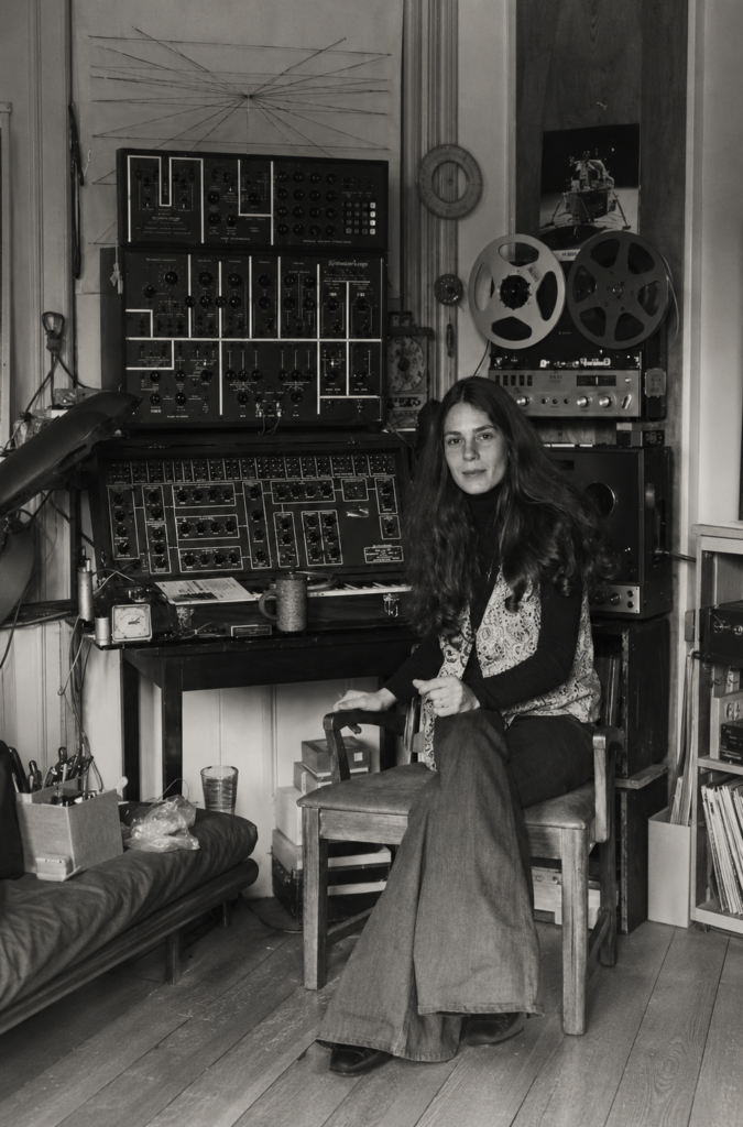 Fotografía en blanco y negro de la compositora e informática Laurie Spiegel sentada en su estudio loft de Nueva York rodeada de sintetizadores analógicos Electrocomp y grabadoras de cinta de bobina abierta.