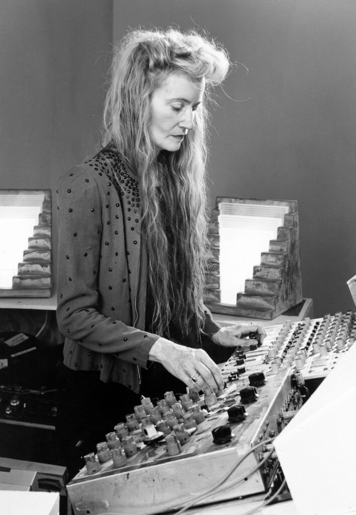 Fotografía en blanco y negro de la compositora e investigadora acústica estadounidense Maryanne Amacher operando una consola de mezcla de audio analógica junto a estructuras de dispersión sónica.