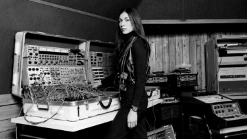 "Sisters with Transistors": las diez pioneras que cambiaron la música electrónica Fotografía antigua en blanco y negro de la compositora Suzanne Ciani en su estudio posando junto a un sistema de sintetizadores modulares portátiles duales Buchla 200 repleto de cables de parcheo.