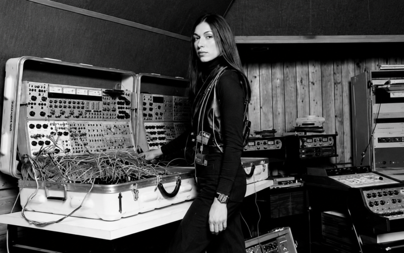 Fotografía antigua en blanco y negro de la compositora Suzanne Ciani en su estudio posando junto a un sistema de sintetizadores modulares portátiles duales Buchla 200 repleto de cables de parcheo.