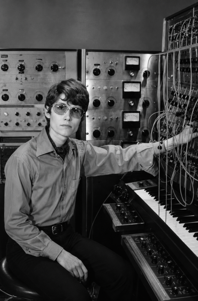Fotografía en blanco y negro de la compositora Wendy Carlos sentada frente a un monumental sintetizador modular Moog con cables de parcheo y un teclado musical en su estudio.