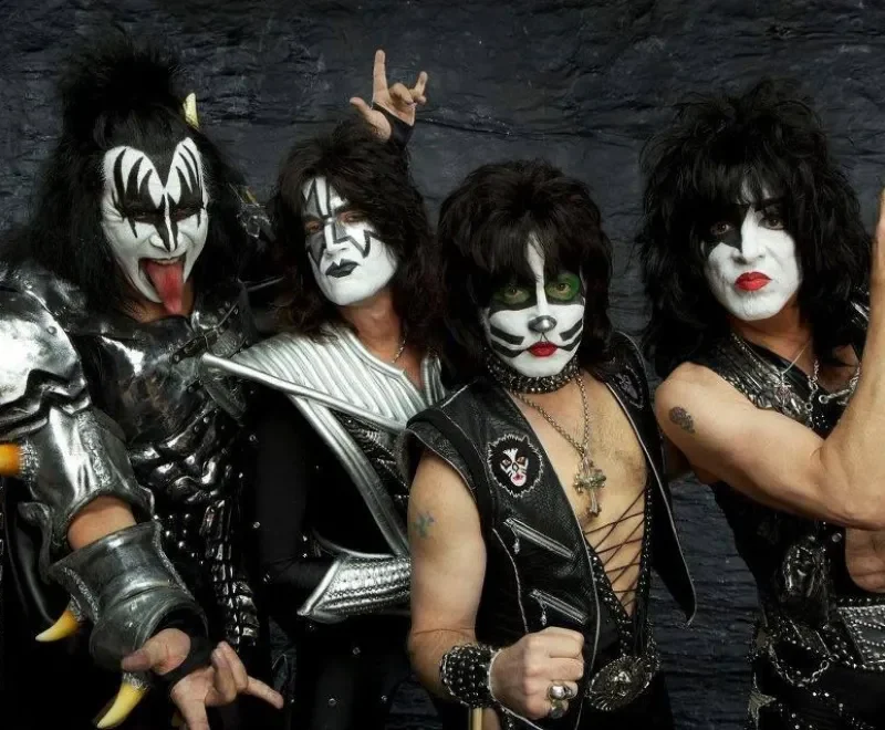 Miembros de la banda estadounidense de rock KISS posando con sus icónicos maquillajes faciales de personajes en blanco y negro frente a un fondo texturizado oscuro. De izquierda a derecha: Gene Simmons como The Demon, Tommy Thayer como The Spaceman, Eric Singer como The Catman y Paul Stanley como The Starchild, mostrando la influencia de la estética teatral en el rock y el posterior corpse paint del black metal.