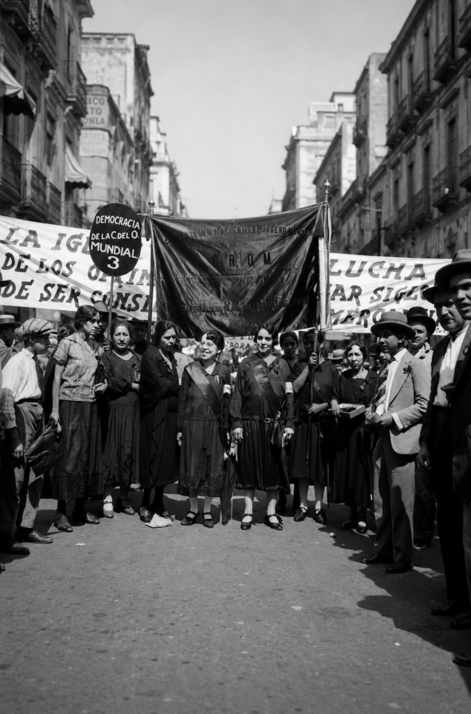 Fotografía histórica en blanco y negro de una manifestación sufragista mexicana en los años 1930; un grupo de mujeres encabeza la marcha sosteniendo estandartes y pancartas que exigen la igualdad de derechos y la democracia mundial.