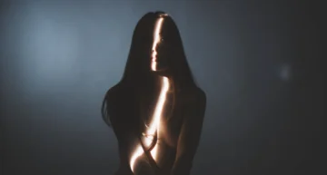 Retrato conceptual y artístico en penumbras, con una franja vertical de luz proyectada sobre su rostro y cuerpo en un fondo gris difuminado.