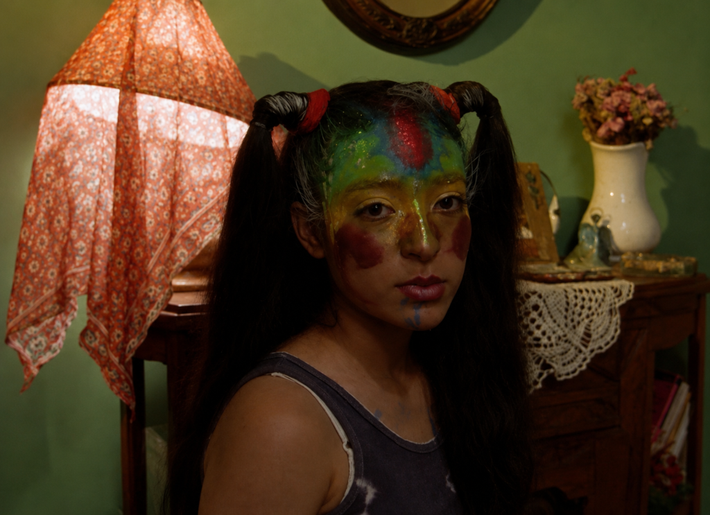 Primer plano de la actriz mexicana Ximena Ayala interpretando a Yessica en 'Perfume de violetas (nadie te oye)' de Maryse Sistach, mostrando el rostro cubierto con un maquillaje de colores llamativos y purpurina en una habitación con paredes verdes.