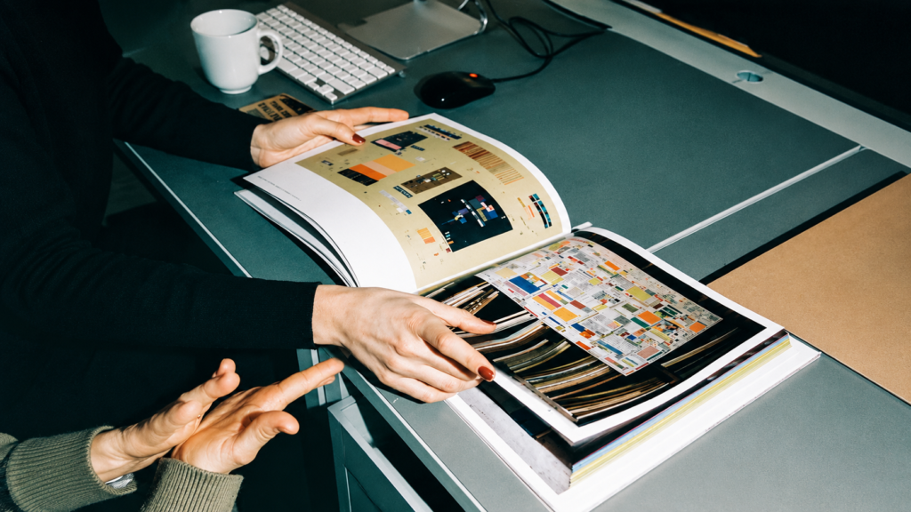 Primer plano de dos personas revisando un libro de arte y diseño editorial abierto sobre un escritorio gris moderno. En el fondo desenfocado se aprecian un teclado de computadora blanco Apple, un ratón negro y una taza blanca de café.