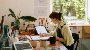 Descarga gratis 3 mil libros de arte en PDF Una ilustradora con pendientes amarillos en forma de flor dibuja digitalmente con un lápiz óptico en una tableta Apple iPad en su taller. Sobre la mesa de madera hay pinceles, botes de cerámica, pinturas de colores y el libro de arte "Monet" de la editorial TASCHEN, cerca de una ventana con plantas en el fondo.