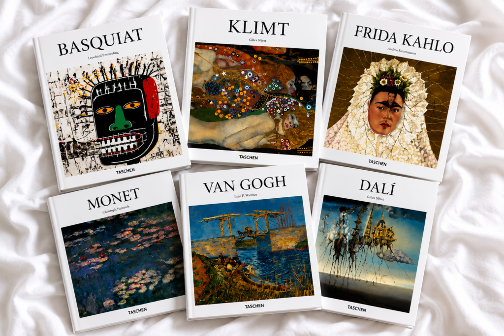 Vista aérea de seis libros de arte de la editorial TASCHEN dispuestos sobre una sábana blanca arrugada. Los títulos corresponden a Basquiat, Klimt, Frida Kahlo, Monet, Van Gogh y Dalí, cada uno mostrando una obra icónica del artista en su portada.