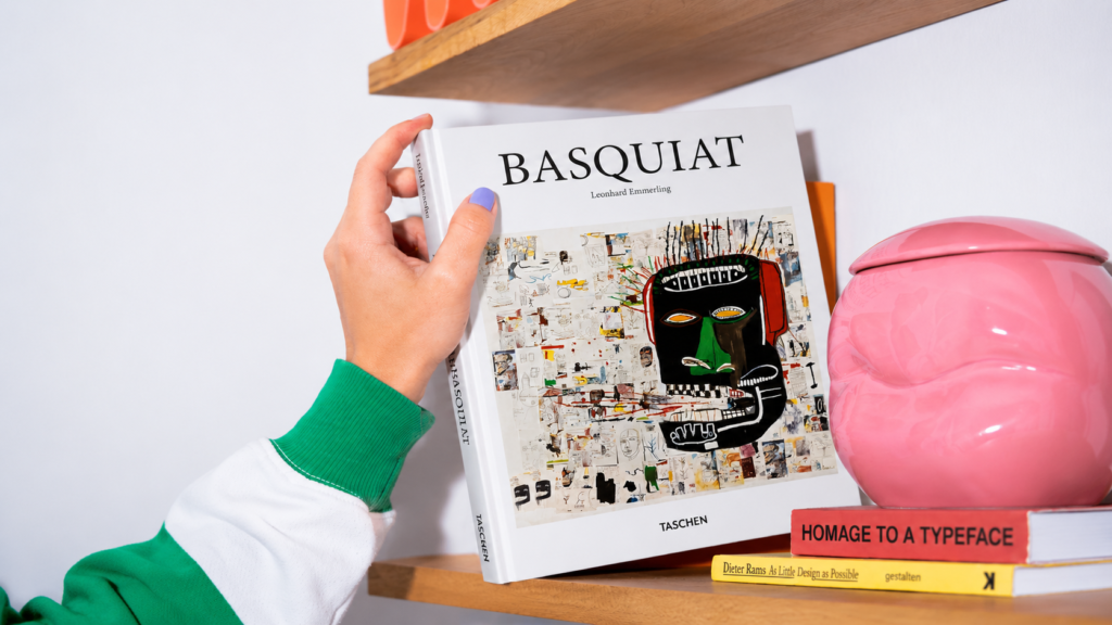 Primer plano de una persona vistiendo una sudadera verde que saca de un estante de madera el libro de arte "Basquiat" de Leonhard Emmerling, editado por TASCHEN. Al lado se aprecia un jarrón rosa brillante y dos libros titulados "Homage to a Typeface" y "Dieter Rams: As Little Design as Possible".