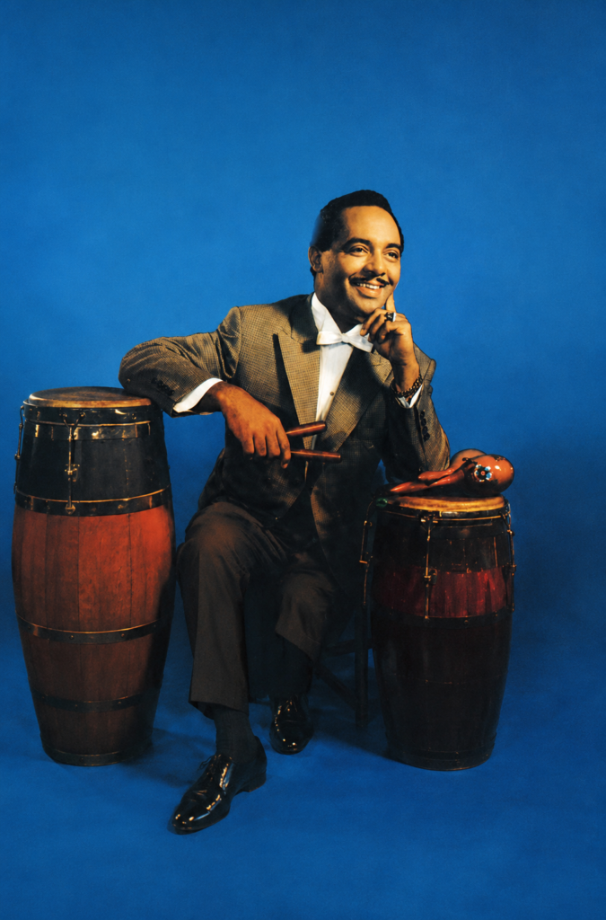 El músico y director de orquesta cubano-mexicano Dámaso Pérez Prado, conocido como el Rey del Mambo, posando sonriente con un traje gris, baquetas en mano, junto a un par de congas y maracas sobre un fondo azul.