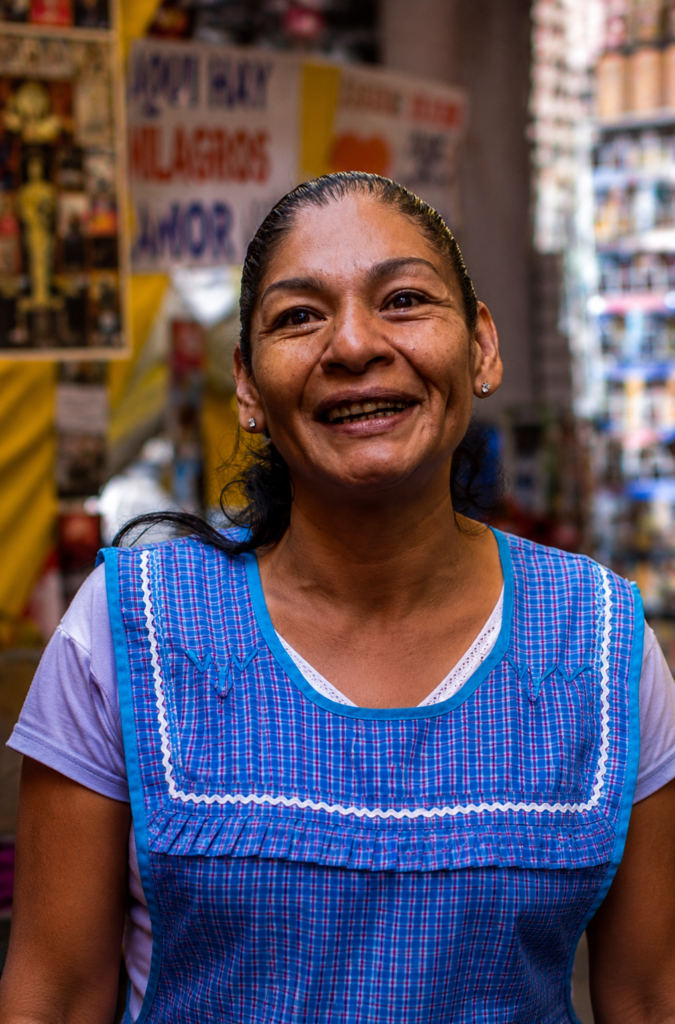 Retrato en primer plano de Lourdes Ruiz Baltazar, conocida como La Reina del Albur de Tepito, sonriendo con un mandil a cuadros azul en su puesto del mercado en la Ciudad de México.