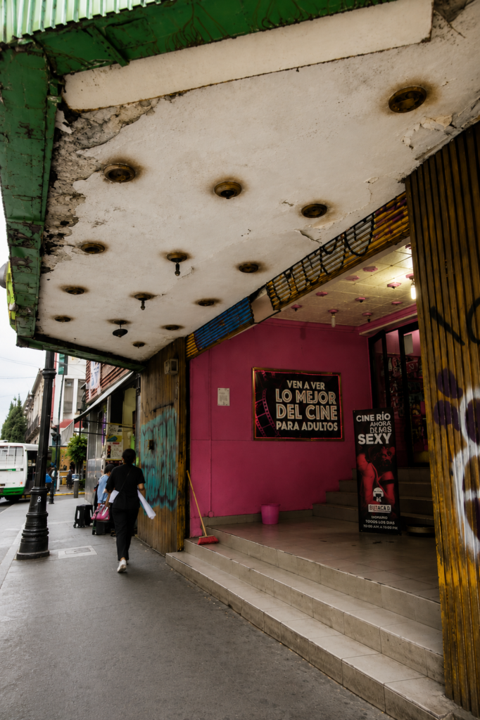 Entrada del Cinema Río en la Ciudad de México con una pared rosa brillante que exhibe un letrero negro con la frase Ven a ver lo mejor del cine para adultos y un cartel vertical que dice Cine Río ahora de lasis sexy, bajo un techo desgastado con sockets vacíos.