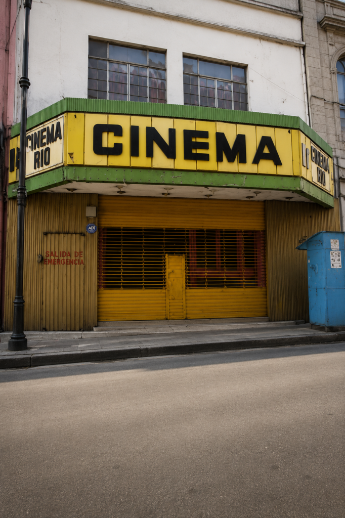 Fachada urbana del Cinema Río en la Ciudad de México antes de su remodelación, mostrando su marquesina amarilla con bordes verdes, muros de lámina acanalada y personas caminando en la acera.