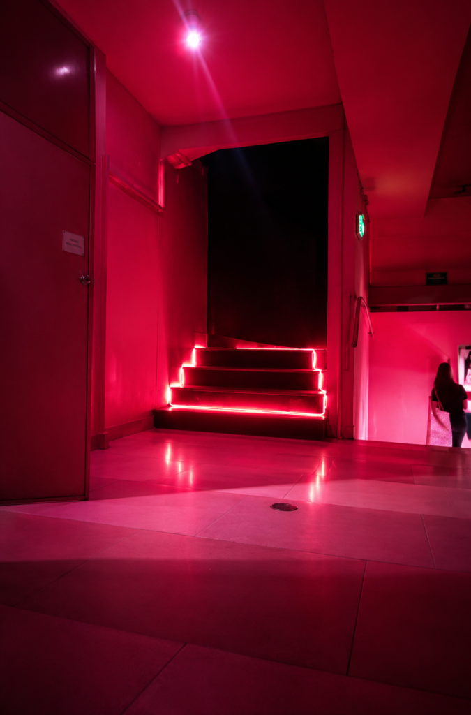 Cabinas privadas del Cinema Río iluminado por una intensa luz rosa y roja, mostrando un pasillo de pisos claros, una puerta a la izquierda y escaleras al fondo delineadas con tiras de luces neón rojas brillantes que suben hacia la penumbra.