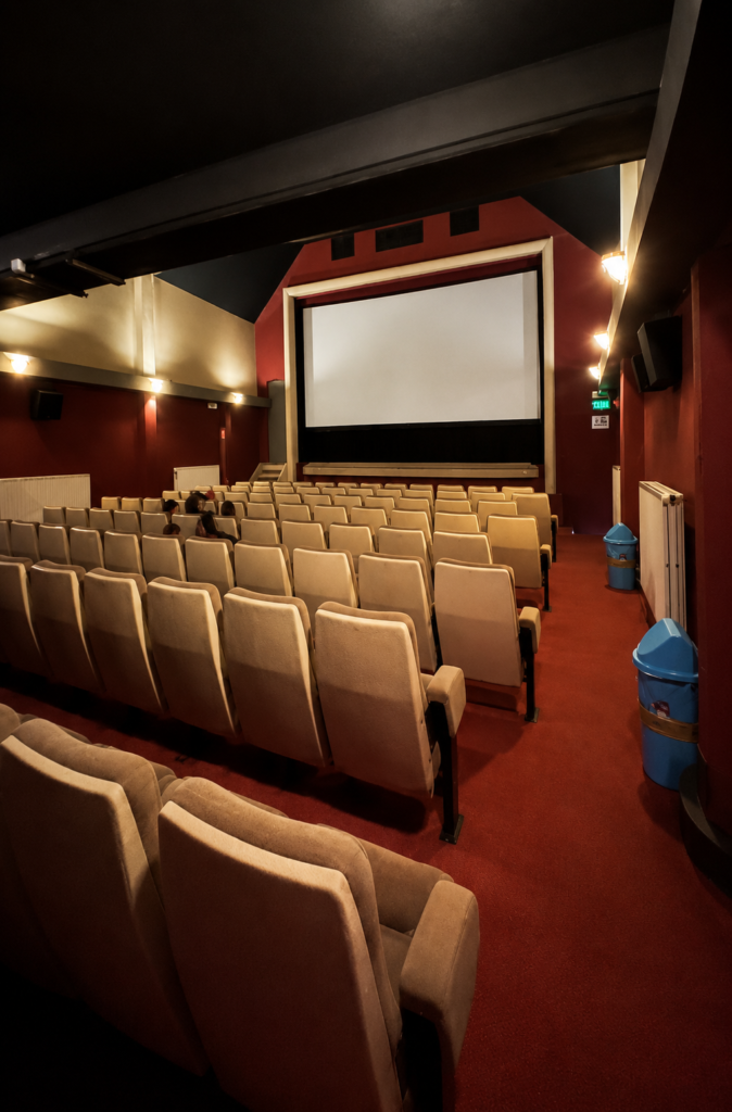 Interior de una sala de cine vacía con filas de butacas de tela color beige, alfombra roja, paredes de tono guinda y una pantalla en blanco al fondo con iluminación tenue.