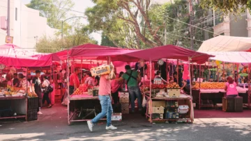 Perspectiva de un tianguis tradicional en la Ciudad de México con puestos bajo lonas rosas y gente realizando intercambios comerciales en el asfalto.