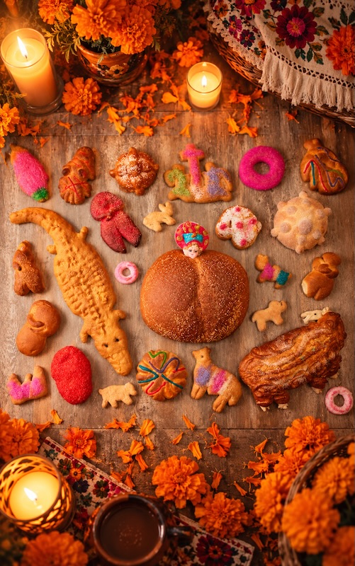 Variedades de pan de muerto con formas humanas, animales y roscas, sobre una mesa con cempasúchiles y velas encendidas.