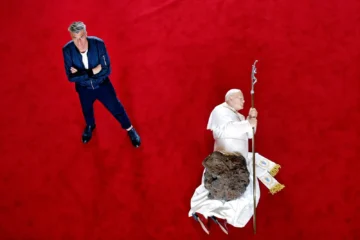 Retrato cenital del artista Maurizio Cattelan parado junto a su famosa escultura del Papa Juan Pablo II derribado por un meteorito sobre una alfombra roja.