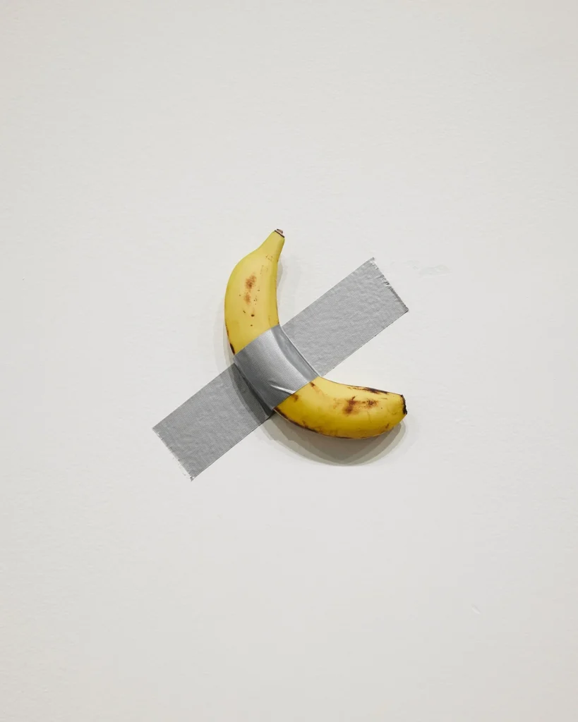 Un plátano maduro adherido de forma diagonal a una pared blanca con un trozo de cinta adhesiva gris, correspondiente a la obra Comedian de Maurizio Cattelan.