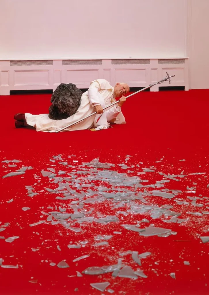 Escultura hiperrealista del Papa Juan Pablo II derribado por un meteorito sobre una alfombra roja con cristales rotos, correspondiente a la obra La Nona Ora de Maurizio Cattelan.