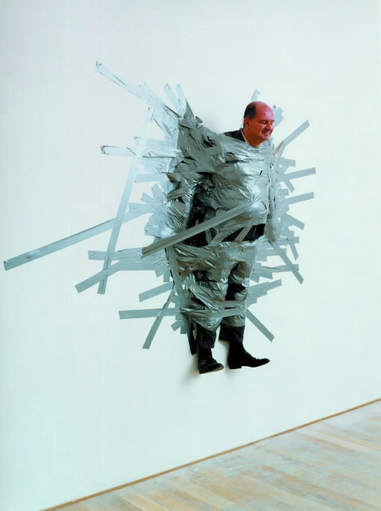 El galerista Massimo De Carlo pegado a la pared con cinta adhesiva gris para la obra A Perfect Day de Maurizio Cattelan.