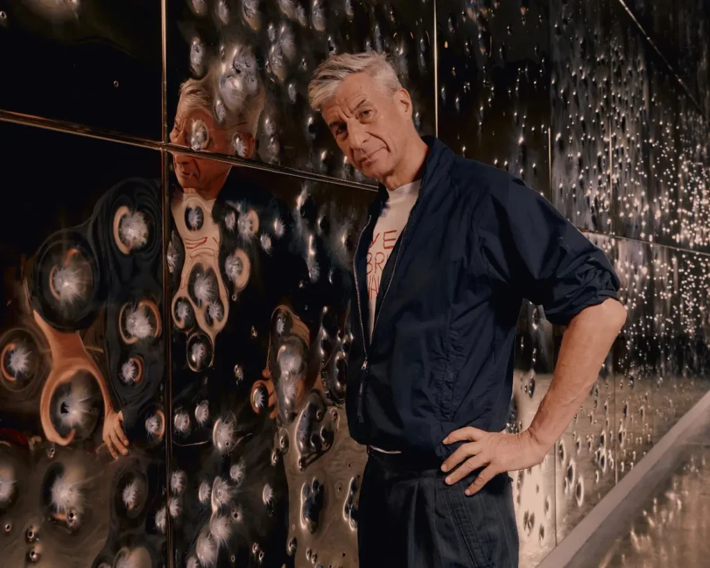 Retrato del artista Maurizio Cattelan posando de perfil junto a su obra de arte titulada MOUTH, consistente en un panel metálico cubierto de oro con perforaciones de impactos de bala reales.