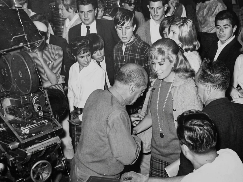 Luis Buñuel dirigiendo a Silvia Pinal en el set de Simón del desierto (1965), rodeados por el equipo de producción y una cámara de cine clásica durante el rodaje del mediometraje.