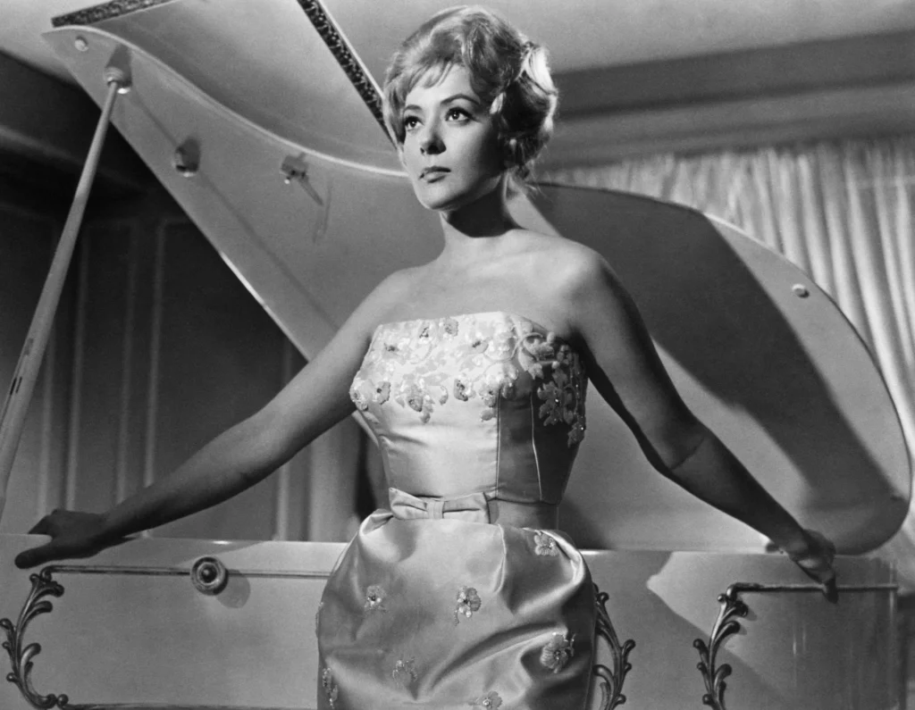 Silvia Pinal interpretando a Leticia 'La Walkiria' en El ángel exterminador (1962), posando con elegancia frente a un piano de cola en una mansión de lujo antes del colapso social de los personajes.