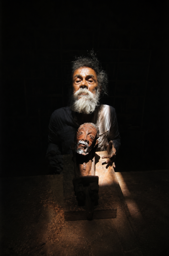 El artista mexicano Francisco Toledo sosteniendo un busto autorretrato de cerámica en claroscuro.