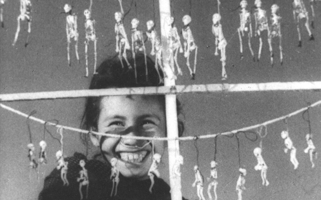 Niña indígena sonriendo en un mercado del Día de Muertos rodeada de juguetes tradicionales de esqueletos colgando, fotograma de la película ¡Que viva México! de Serguéi Eisenstein.