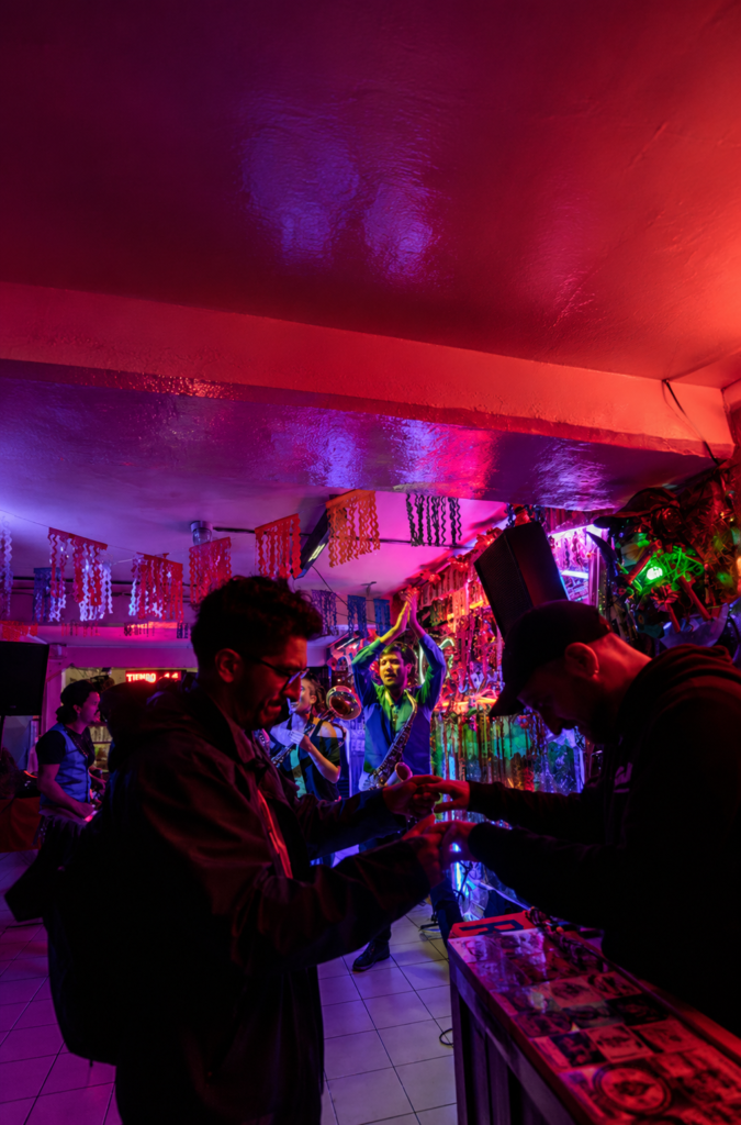Asistentes bailan alegremente en primer plano bajo una iluminación de luces de neón en tonos magenta, rosa y morado, mientras un músico toca el saxofón sobre el escenario del restaurante y marisquería psicodélica La Caña en la Colonia Doctores de la Ciudad de México.