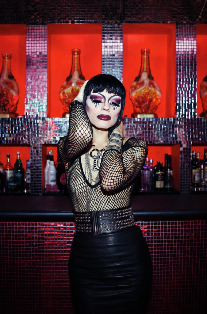 Una drag queen con un elaborado maquillaje artístico en tonos rojos, negros y blancos posa de manera expresiva frente a la barra del icónico bar queer La Purísima en la Ciudad de México, decorado al fondo con estatuillas religiosas kitsch iluminadas en tonos naranja y rojo.