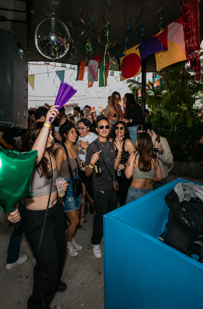Un grupo de jóvenes sonríe, platica y sostiene bebidas en la concurrida terraza al aire libre de Revuelta Queer House en la Colonia Roma de la Ciudad de México, bajo un techo decorado con guirnaldas y banderines multicolores de la diversidad.