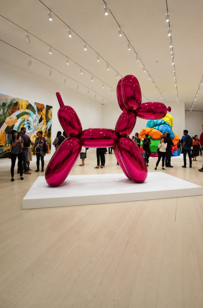 Escultura gigante de acero inoxidable Balloon Dog Magenta de Jeff Koons sobre una plataforma blanca en la sala principal del Museo Jumex, con visitantes observando la obra y pinturas contemporáneas de fondo en la Ciudad de México.