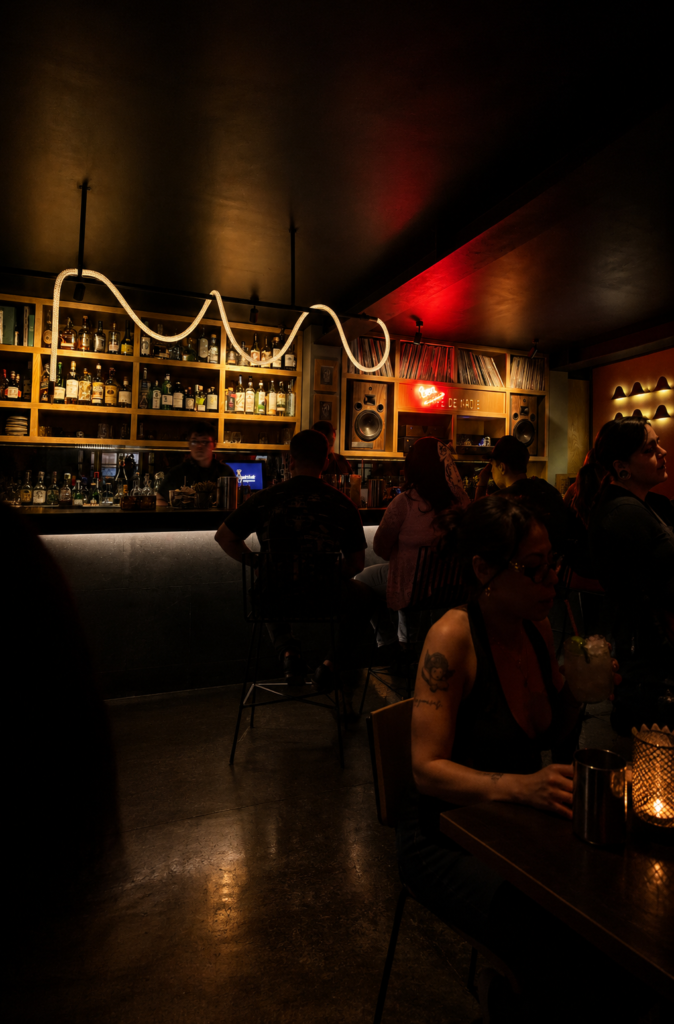 Clientes sentados en la barra iluminada de Café de Nadie Listening Bar en la CDMX, con DJs al fondo, un letrero de neón rojo y estanterías llenas de discos de vinilo.